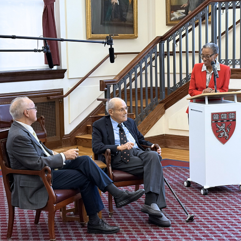 Joan Reede stands behind an HMS-branded lectern while George Daley and David G. Nathan sit in two chairs in the Waterhouse Room.