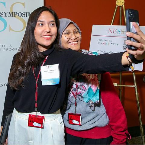 Two young women, one with long dark hair and the other in a hijab, take a selfie in front of a NESS banner.