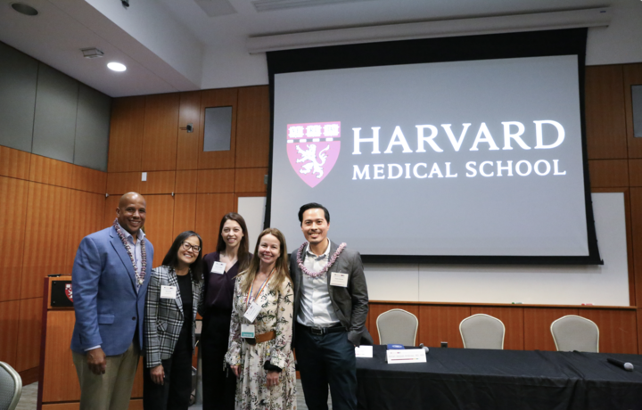 Four panelists and the moderator for the mental health forum stand in front of a projector with the HMS logo.  