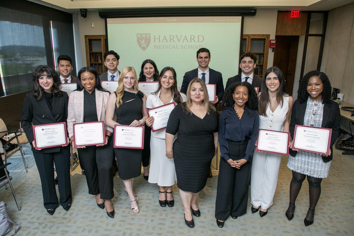 Eleven VRIP students pose with their certificates alongside two staff members. The Harvard Medical School logo is projected on a screen behind them. 