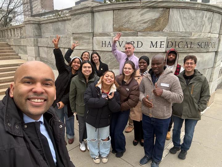 Pablo da Silva takes a selfie in front of the Harvard Medical School sign on the Quad. He is standing with a BHCC professor and 10 BHCC students, some of whom are throwing peace signs or waving.