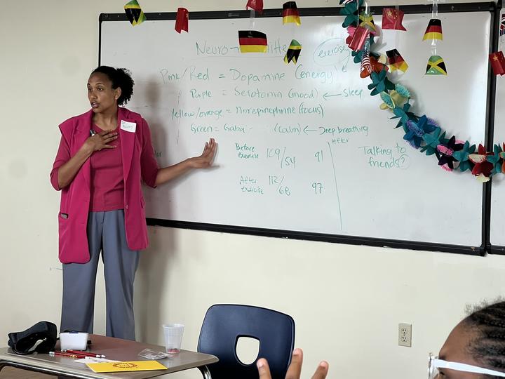 Frinny Polanco is gesturing to herself and a whiteboard with neurotransmitter terms on it.
