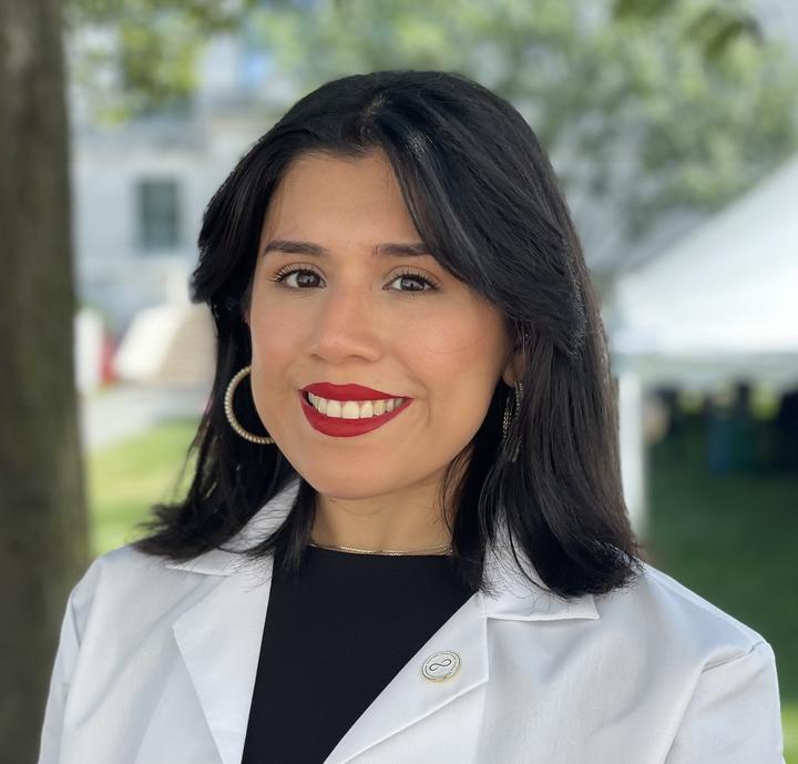 Genesis Perez-Melara is wearing a black blouse and white coat. She is standing outside in front of a tree.