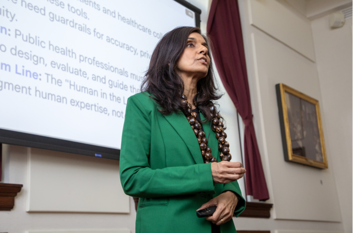 Monica Bharel is wearing a green blazer and necklace made of kukui nuts. She is standing in front of a projector screen in the Waterhouse Room and speaking.