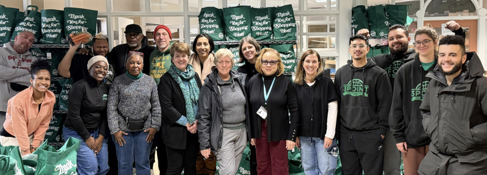 The Dimock Center and OCCE crew stands in front of green reusable bags of packaged foods. The bags say, "Thank You For Shopping Here!"
