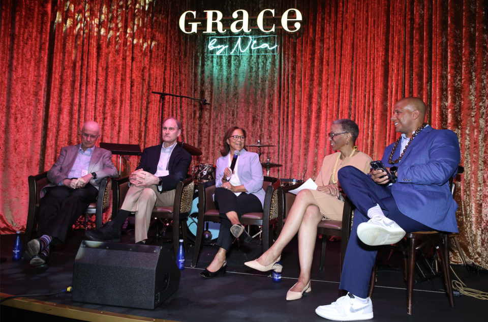 A panel of people sit on stage chatting in the Grace by Nia venue. There is a red curtain and neon sign in the background.