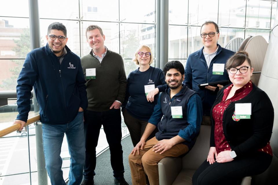 A group of six people, all standing or sitting, pose for a photo in front of the large windows of the Vertias Science Center.