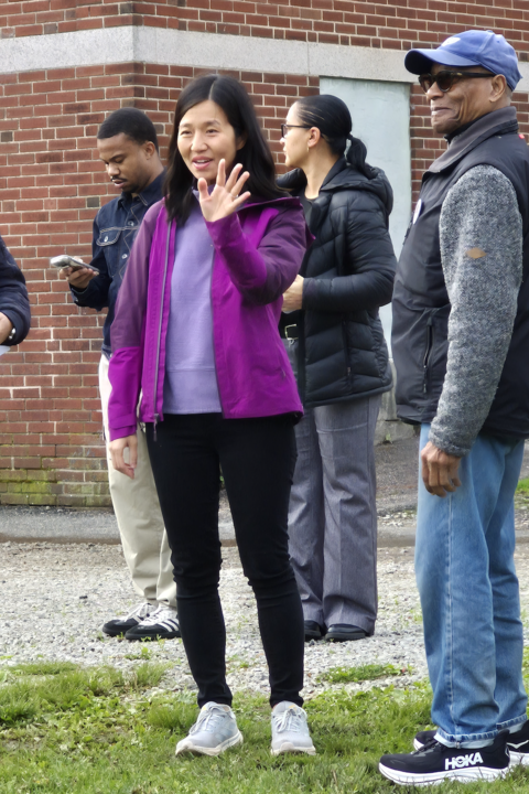 Boston Mayor Michelle Wu waves at someone while at the Mother's Day Walk. She is wearing black leggings, a lavender top, and purple jacket. There are a few people standing around her.