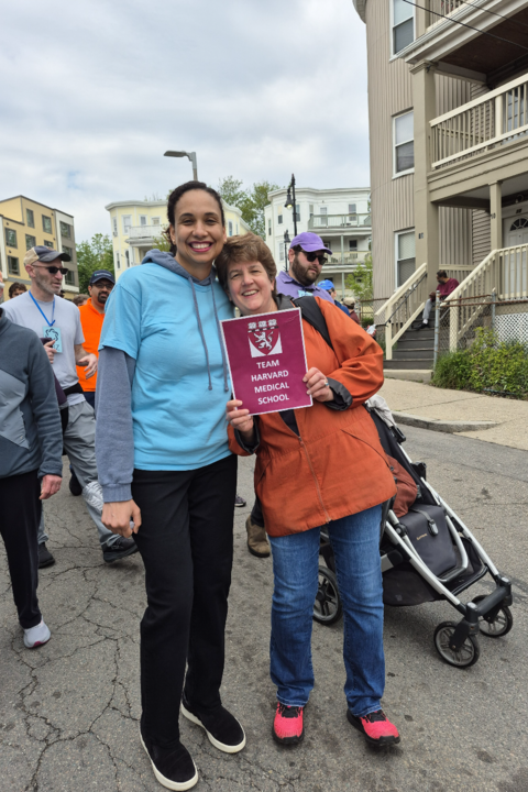 Two members of the OCCE staff stand outside during the Mother's Day Walk. One of them is holding up a red Team Harvard Medical School sign.