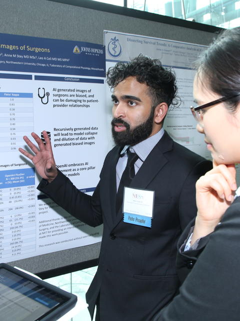 A mean with black hair and facial hair gestures to a poster beside him while two people listen to him talk.