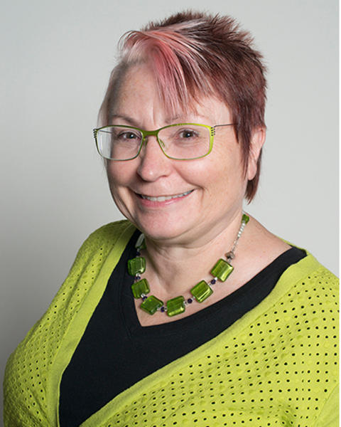 Rebecca Ward is wearing a bright green cardigan, black shirt, and beaded green necklace. Her glasses frames are also bright green.