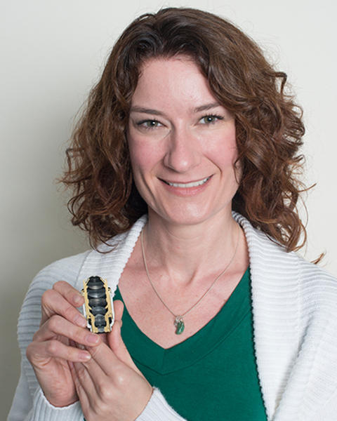 Jennifer Waters is holding up a split section of an objective lens. She is wearing a green shirt and white cardigan.