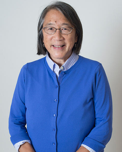 Beverly Woo is wearing a blue cardigan and blue striped shirt. Her hands are clasped in front of her.