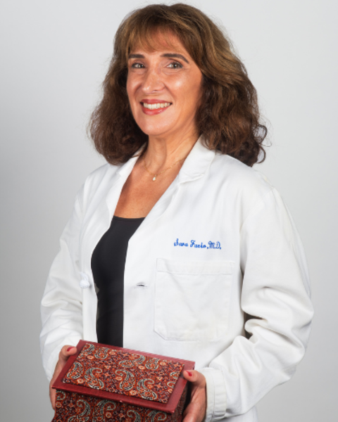 Sara Fzio is wearing a white doctor's coat with her name embroidered on it. She is holding a patterned red wooden box.