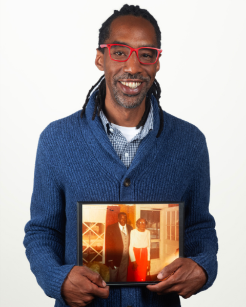 Christopher Crosby is wearing a blue knit cardigan and bright red glasses. He is holding a sepia-toned photo of his great grandfather.