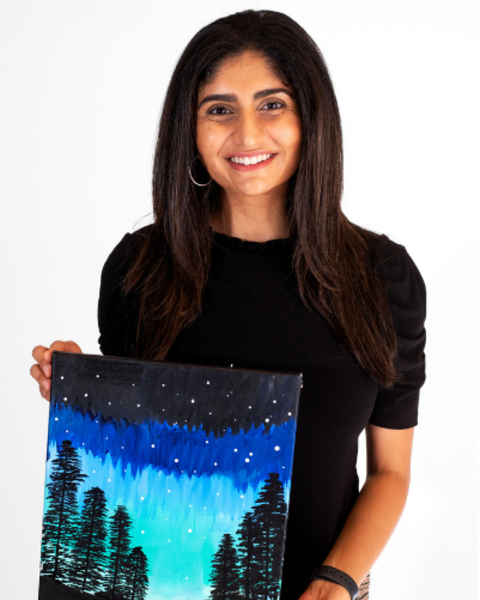 Mansi Totwani is holding a painting of trees with a blue aurora in the night sky.