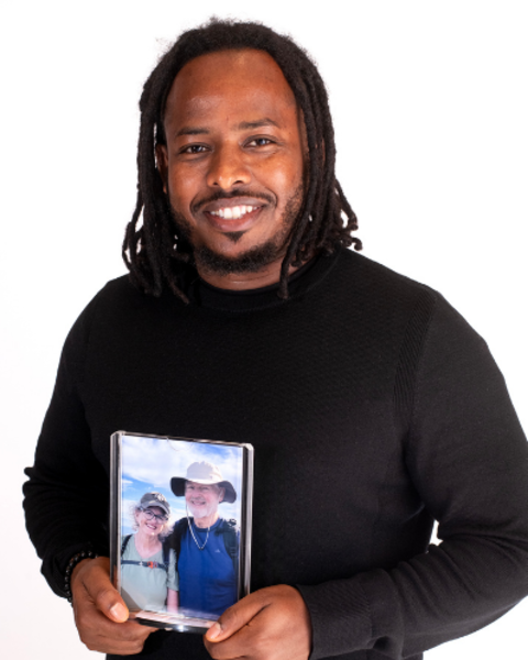 Merhawi Wells-Bogue is holding up a framed photo of his parents. He is wearing a black long sleeve shirt.