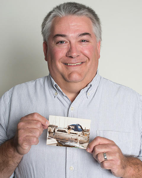 Philip Benware is wearing a blue striped button-down shirt and holding up a photo of his grandfather on a boat.