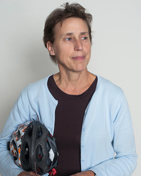 Constance Cepko is wearing a dark shirt and light blue cardigan. She is looking towards her left and is holding a bike helment and glasses.