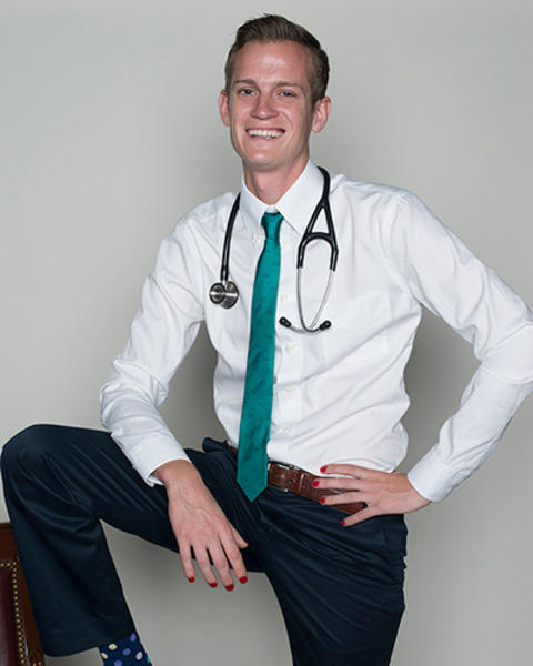 Cary Crall has one foot on a chair armrest with one hand resting on that lifted leg and his other hand on his hip. He is wearing bright red nail polish and has colorful blue polka dotted socks.