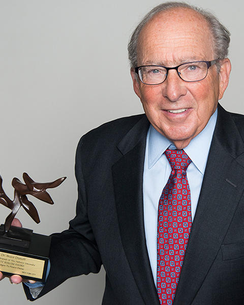 Bruce Donoff is holding up a carved tree of peace sculpture on a black stand. He is wearing a black blazer, light blue shirt, and red patterned tie.