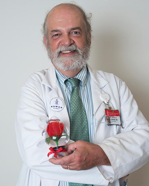 Lachlan Forrow is holding up a red rose preserved in glass. He is wearing a white doctor's coat, striped blue shirt, and green patterned tie.