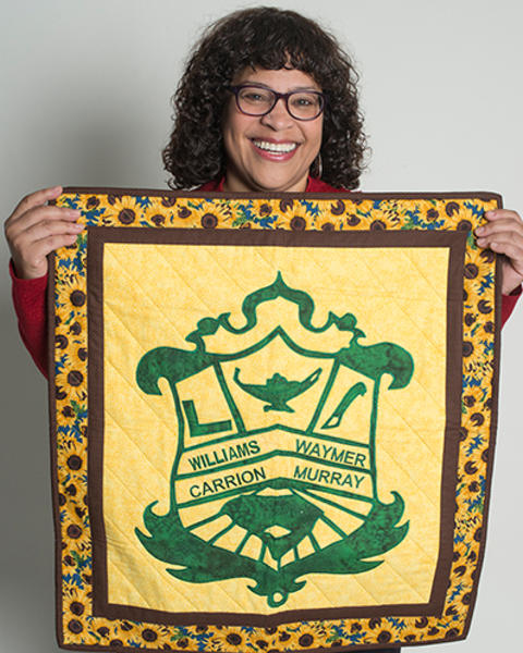 Yvonne Gomez-Carrion is holding up a yellow, brown, and green quilt with her family's coat of arms.