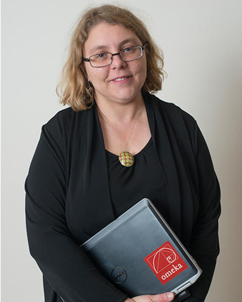 Emily Novak Gustainis is holding her laptop in front of her. She is wearing a black shirt, black cardigan, and green patterned necklace.
