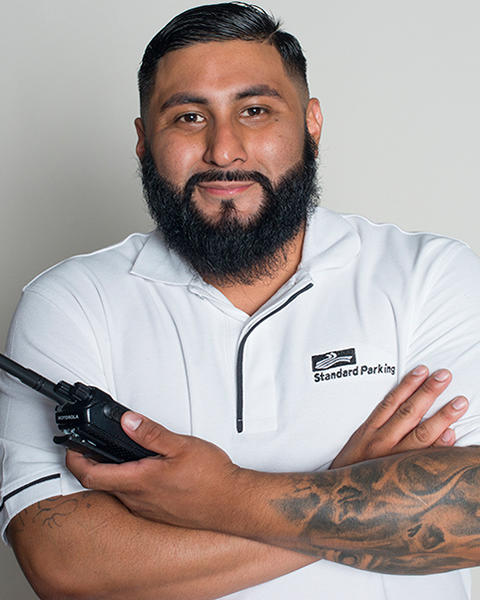 Simon Ibanez has his arms crossed in front of him and is holding a walkie-talkie. He is wearing a white polo.