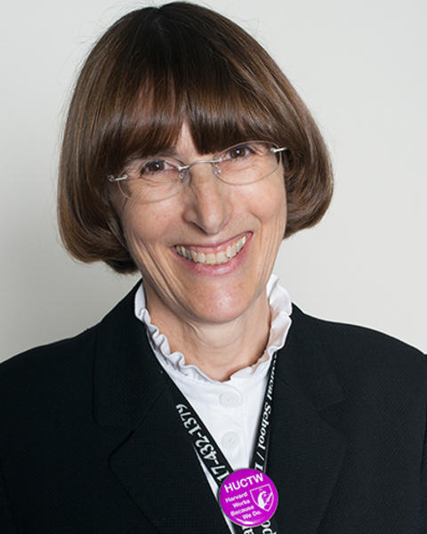 Maura Kilcommons is wearing a black blazer and white blouse with a ruffled collar. She is a purple HUCTW pin on her black Harvard Longwood Campus lanyard.