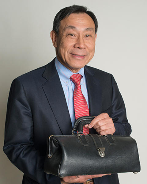 George King is holding up a small black doctor bag. He is wearing a blue blazer, gray pants, blue shirt, and red patterned tie.