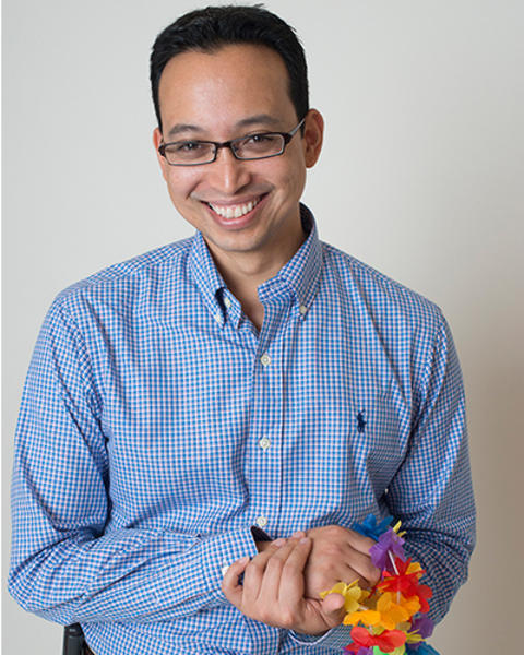 Robert Kitts is holding a rainbow lei. He is wearing a plaid blue shirt and khakis.