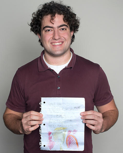 Mounir Koussa is holding a piece of notebook paper with a drawing from his sister. He is wearing a maroon polo and jeans.
