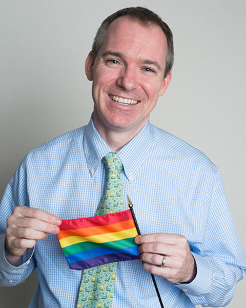 Graham McMahon is wearing blue shirt and green and yellow tie. He is holding a small rainbow flag.
