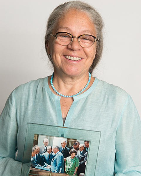 Nancy Oriol is wearing a light blue blouse and turquoise beaded necklance. She is holding a framed photo of the Head Start Program signing ceremony in the Rose Garden.