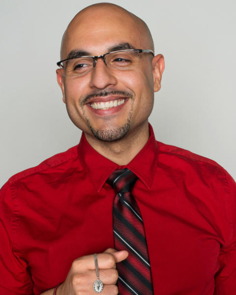 Numa Perez is holding a little silver medal of the Virgin Mary. He is wearing a red shirt and red, black, and white striped tie.