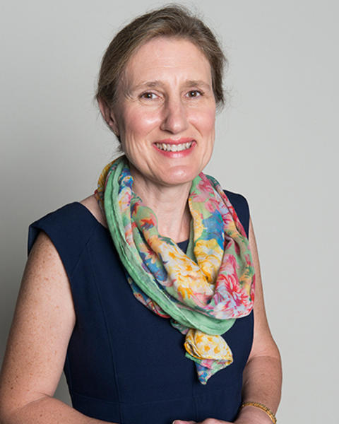 Jane Barrow is wearing a colorful sarf and dark blue dress. Her hands are clasped in front of her. 