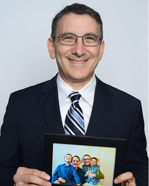 Mark Schuster is wearing a dark striped blazer, white shirt, and blue striped tie. He is holding a framed photo of his family. 