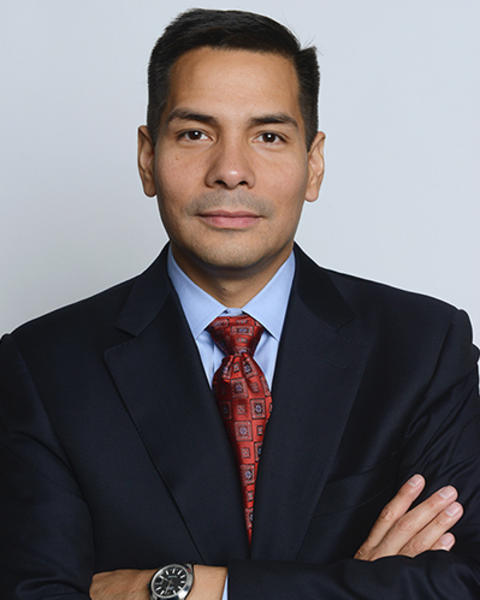 Thomas Sequist is wearing a dark blazer, blue shirt, and red patterned tie. He has his arms crossed in front of him.
