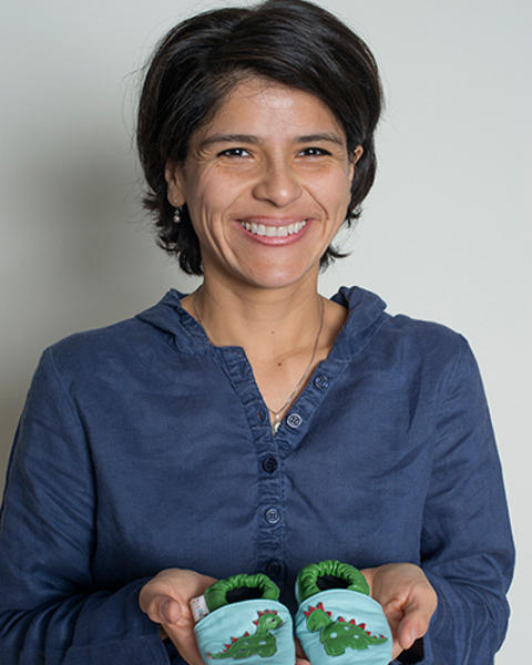 Mercedes Becerra is holding up a pair of blue and green baby shoes with dinosaurs on them.