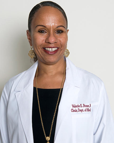 Valerie Stone is wearing a black shirt under her white coat that has her name embroidered on it. She is also wearing gold earrings and pendant necklace.
