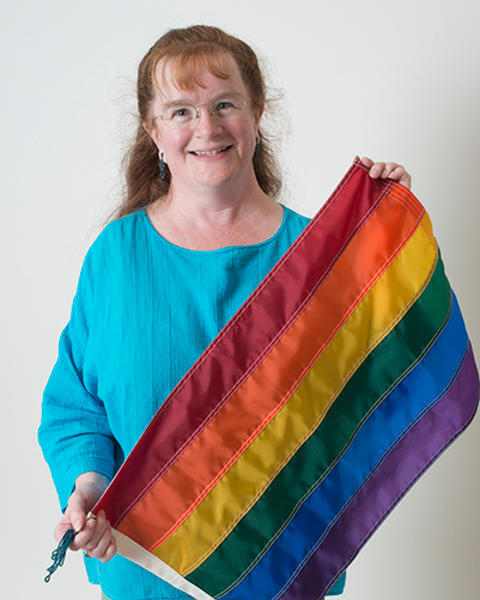 Barbara Sweeny is wearing a turquoise shirt and green pants. She is holding up a rainbow flag.