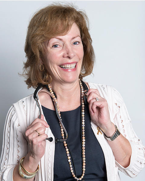 Nancy Tarbell is wearing a dark top and white cardigan. She also has a beaded necklace, lanyard, and stethoscope around her neck.