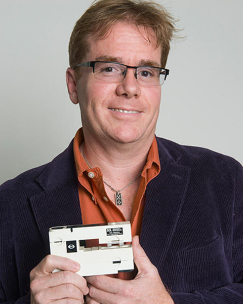 David Van Vactor is holding a small white microscope. He is wearing a dark corduroy blazer and orange shirt.