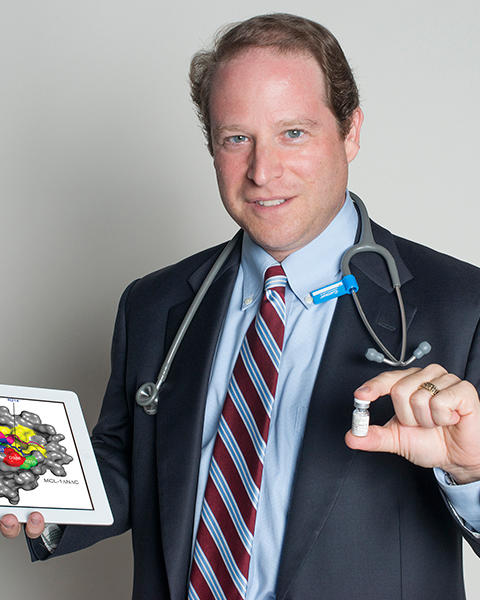 Loren Walensky is holding a vial in his left hand and an image of a structure of a biochemical complex in his right hand.