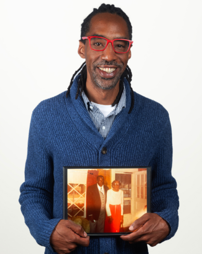 Christopher Crosby is wearing a blue knit cardigan and bright red glasses. He is holding a sepia-toned photo of his great grandfather.