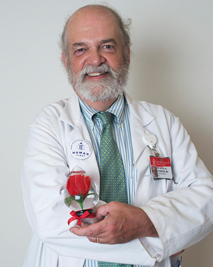 Lachlan Forrow is holding up a red rose preserved in glass. He is wearing a white doctor's coat, striped blue shirt, and green patterned tie.