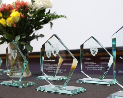 Angular glass awards are spread across a black table. There is a vase of yellow, orange, and white flowers in the background.