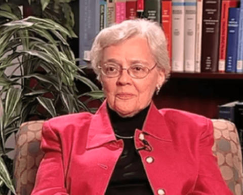 Eleanor Shore is wearing a pink jacket and black top. There is a plant and bookshelf behind her.