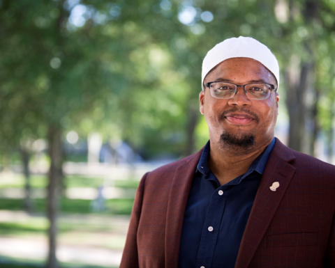 Khalil Abdur-Rashid stands outside by some trees and wears a white kufi, burgundy blazer, and dark blue button down shirt. 